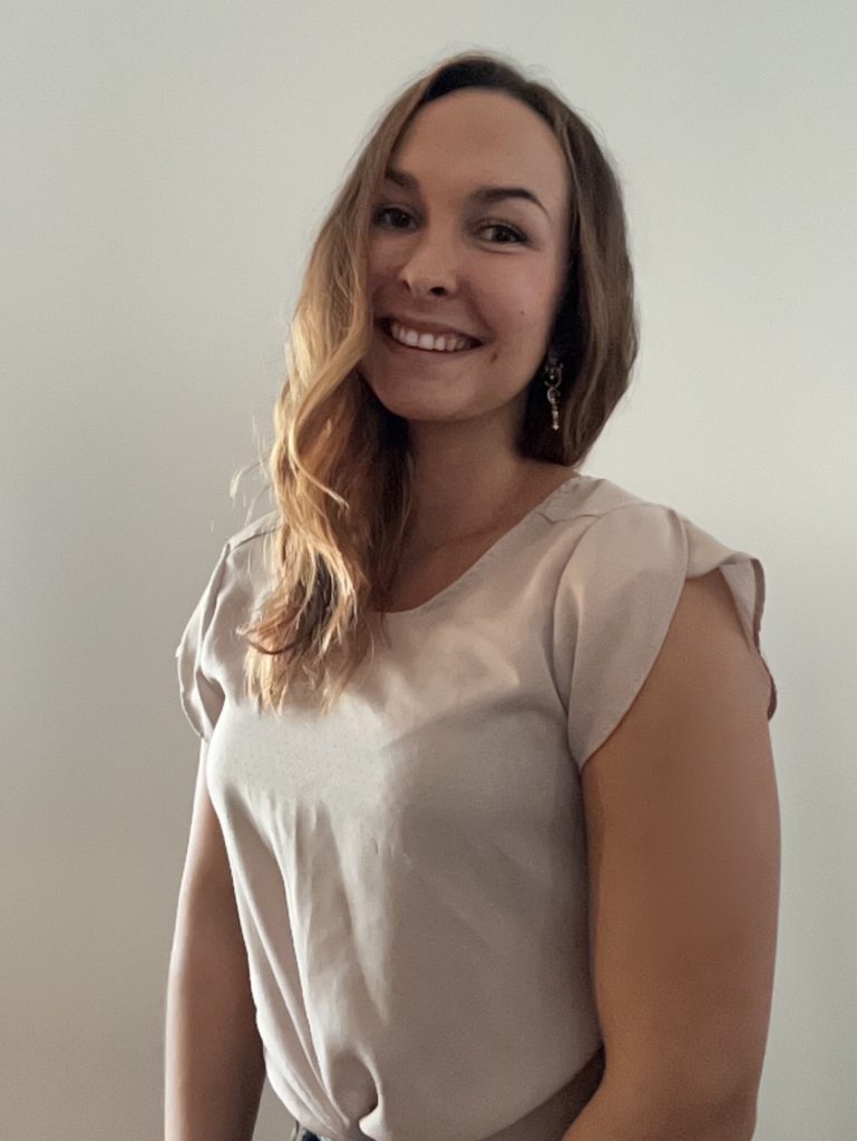 Portrait of a smiling woman with shoulder-length light brown hair, wearing a pale pink blouse, standing against a neutral wall.