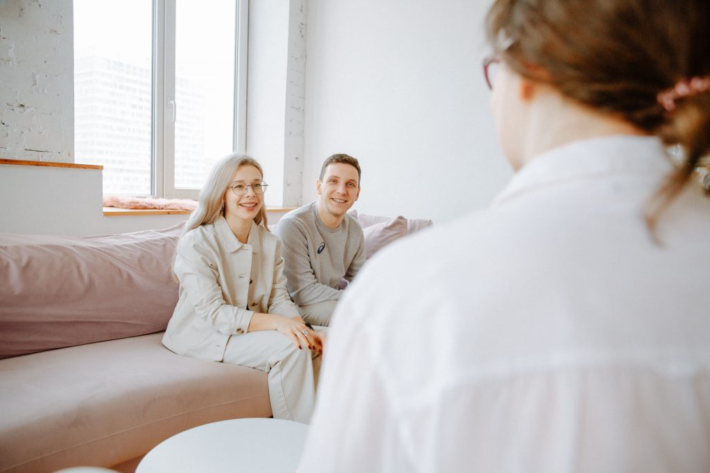 A couple smiles while sitting on a sofa during a counseling session indoors.