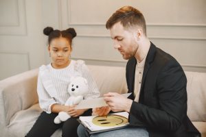 A child holding a plush toy listens to a man in a therapy session.